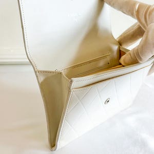 May include: A white leather wallet with a quilted pattern and a snap closure. The interior features a gold-colored lining and a small pocket. The brand name "CHANEL" is visible on the inside flap.
