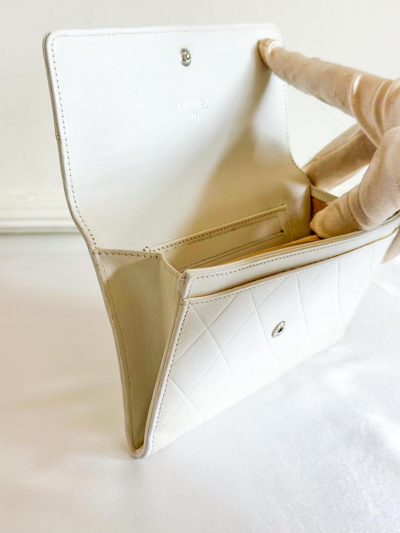 May include: A white leather wallet with a quilted pattern and a snap closure. The interior features a gold-colored lining and a small pocket. The brand name "CHANEL" is visible on the inside flap.
