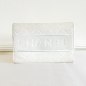 May include: A white quilted leather wallet with the brand name "CHANEL" embossed on the front. The wallet has a rectangular shape with a flap closure. The overall design is minimalist and elegant.