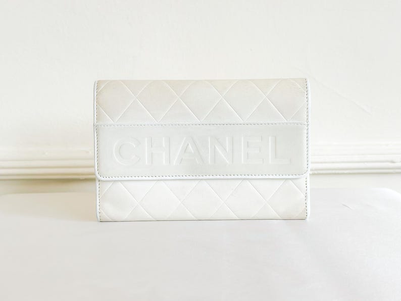 May include: A white quilted leather wallet with the brand name "CHANEL" embossed on the front. The wallet has a rectangular shape with a flap closure. The overall design is minimalist and elegant.