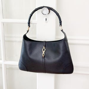May include: Black leather shoulder bag with a bamboo handle. The bag features a gold-tone clasp and hardware. The design is a classic, elegant style, suitable for everyday use. The bag is hanging on a white door.