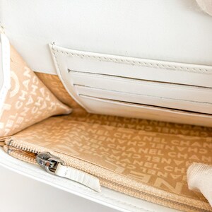 May include: A white leather wallet with a zipper closure and multiple card slots. The interior lining is beige with a repeating logo pattern. The wallet is open, revealing the interior design.