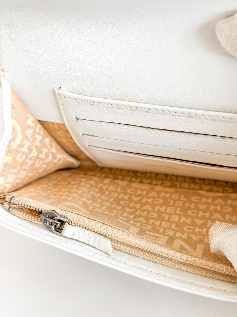May include: A white leather wallet with a zipper closure and multiple card slots. The interior lining is beige with a repeating logo pattern. The wallet is open, revealing the interior design.
