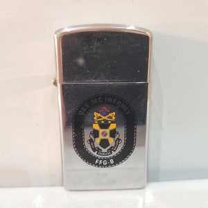 May include: Silver lighter with a black and yellow emblem of the USS McInerney FFG-8. The emblem features a shield with a cross and a crown, surrounded by a rope.