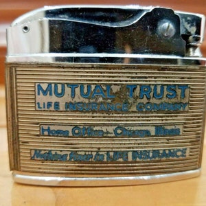 May include: Vintage metal lighter with a silver top and a gold-toned body. The body features the text "MUTUAL TRUST LIFE INSURANCE COMPANY" in blue lettering. The lighter has a rectangular shape.