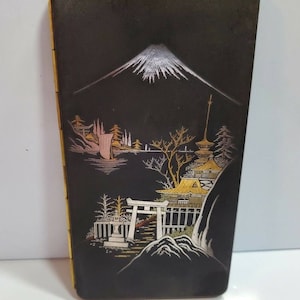 May include: Black cigarette case with a gold and silver Japanese mountain scene. The scene includes a mountain, a waterfall, a pagoda, and a torii gate.