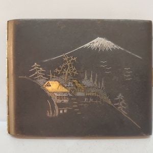 May include: A silver cigarette case with a Japanese mountain scene design. The case is decorated with gold and white accents, depicting a mountain, trees, and a small house.