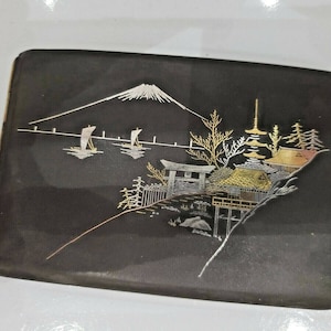 May include: A black lacquered cigarette case with a silver and gold design of a Japanese mountain scene. The scene includes a mountain, a pagoda, a gate, trees, and boats.