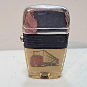 May include: A vintage chrome and gold-tone lighter with a black ribbed centre section. The lighter features a red truck and trailer graphic on the front. The top is chrome, and the bottom is gold-toned.