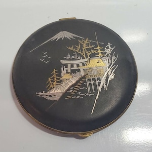 May include: A black compact with a gold and silver design of a Japanese mountain scene. The design includes a mountain, a temple, trees, and birds.