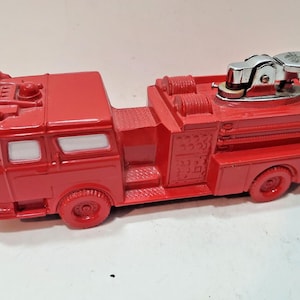 May include: A red fire truck lighter with a silver lighter head. The toy-like lighter has a detailed design, including a ladder, wheels, and windows. The lighter is made of a solid material and is a collectible item.