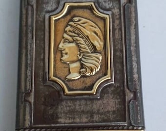 May include: A silver and gold matchbox with a raised image of a woman's profile wearing a crown. The matchbox has a gold trim around the edges.