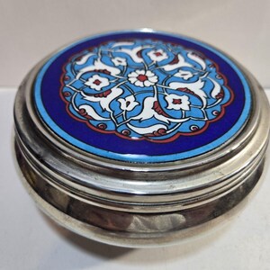 May include: A round, silver-toned trinket box with a detailed, colorful lid. The lid features a vibrant blue background with a floral design in white, light blue, and red. The box is likely made of metal and has a shiny finish.