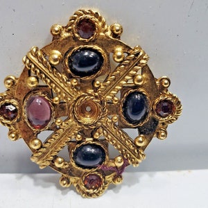 May include: A gold-coloured brooch with a cross-like design. The brooch features oval-shaped, dark-coloured gemstones and smaller, round, red-toned gemstones. The design includes rope-like detailing and small, gold-coloured spheres.