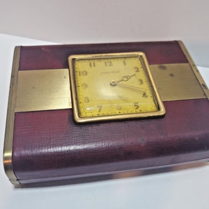 May include: A vintage travel clock with a burgundy leather case and a gold-colored metal frame. The clock face is square with a yellow background and black numerals. The clock is approximately 4 inches wide and 3 inches tall.