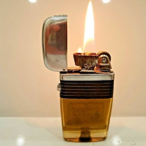May include: A vintage lighter with a gold and black design. The lighter is open and the flame is lit. The lighter has a silver top and a clear plastic body.