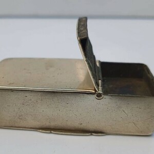 May include: A small, rectangular, silver-colored metal box with a hinged lid. The lid is open, revealing the empty interior of the box.