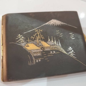 May include: A black cigarette case with a gold and silver design of a Japanese mountain scene with houses and trees.
