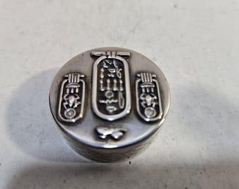 May include: Silver trinket box with an Egyptian hieroglyphic design. The box is round and has a lid with three ankh symbols.