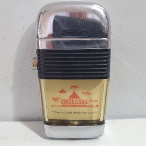 May include: A vintage chrome and gold-coloured Chickasha lighter. The lighter features a black ribbed section and the text "Chickasha" with the phrase "Just A Little Better For Less".