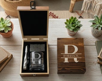 May include: A wooden gift set with a personalized whiskey glass, tongs, and whiskey stones. The glass and wooden box are engraved with the letter "R" and the name "Robertson". The set is displayed open, revealing the contents.