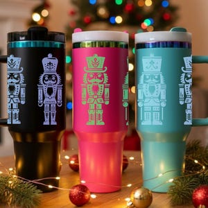 May include: Three insulated travel mugs with handles, each featuring a nutcracker design. The mugs are black, pink, and teal, with the nutcracker designs in contrasting colors. The mugs are on a wooden surface with Christmas decorations.
