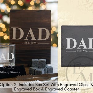 a set of items, including a wooden box with the word "DAD" engraved on it, a glass, and a set of gray stones. The items are arranged on a table, with a blurred background that includes a Christmas tree.