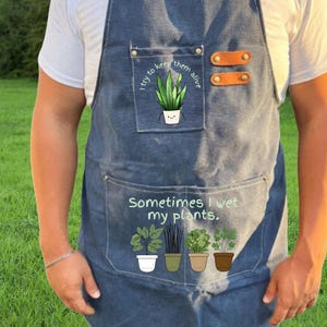 May include: Denim apron with a pocket featuring a potted plant illustration and the text "I try to keep them alive." Below, the apron has the text "Sometimes I wet my plants" and illustrations of potted plants.