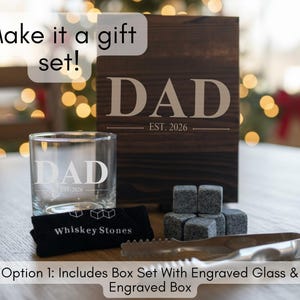 a gift set that includes a wooden box with the word "DAD" engraved on it, a glass, and whiskey stones. The text on the image suggests that the gift set can be customized with an engraved glass and a carved wooden box.