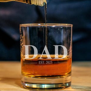 A glass of amber-colored liquid, likely whiskey, with the word "DAD" printed on it, and the text "EST. 2025" below it.