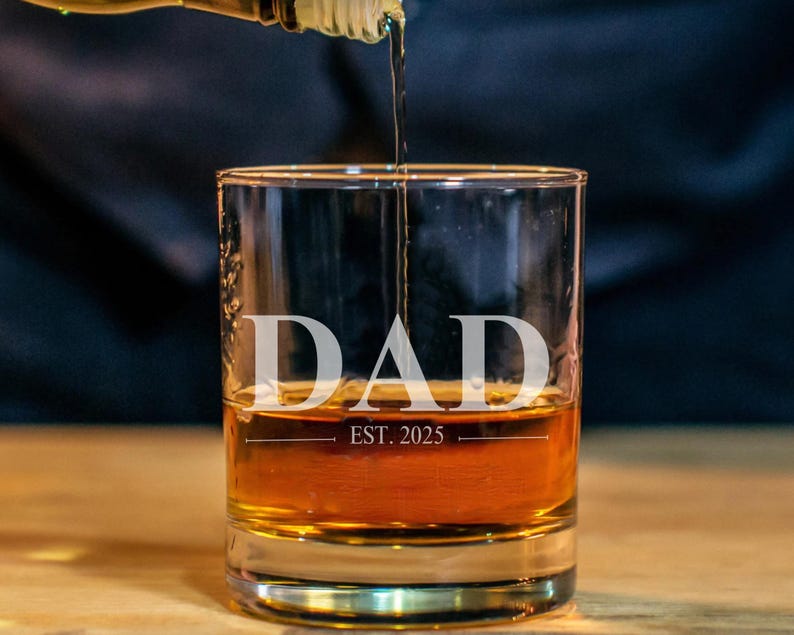 A glass of amber-colored liquid, likely whiskey, with the word "DAD" printed on it, and the text "EST. 2025" below it.