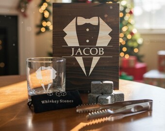 May include: A personalized whiskey gift set featuring a wooden box with a tuxedo design and the name "JACOB". Includes a rocks glass, whiskey stones, a black pouch, and tongs. The set is displayed on a wooden table.