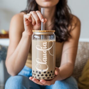 May include: A clear glass boba tea cup with a bamboo lid and a clear straw. The cup is filled with a light brown beverage and boba pearls. The name "Claudia" is written in a cursive font on the side of the cup.