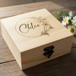 May include: A light-colored wooden box with a floral design and the name "Chloe" engraved on the lid. The box has a dark metal clasp and is sitting on a wooden surface. Daisies and pink flowers are in the background.