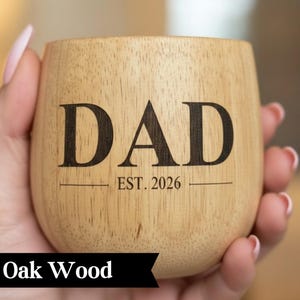 A wooden mug with the text "DAD EST. 2026" printed on it, held in a person's hand.