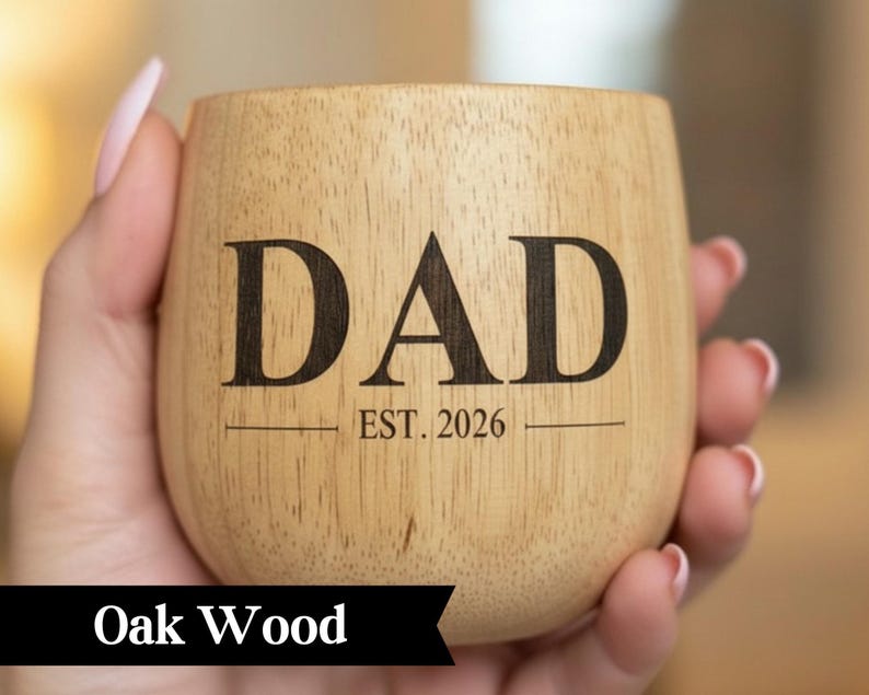A wooden mug with the text "DAD EST. 2026" printed on it, held in a person's hand.