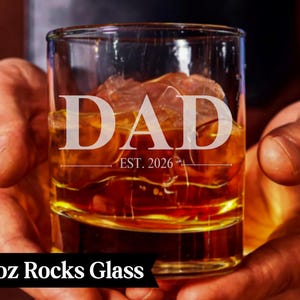 A person's hands holding a 10 oz glass filled with amber-colored liquid, with the word "DAD" and "EST. 1926" engraved on the glass.