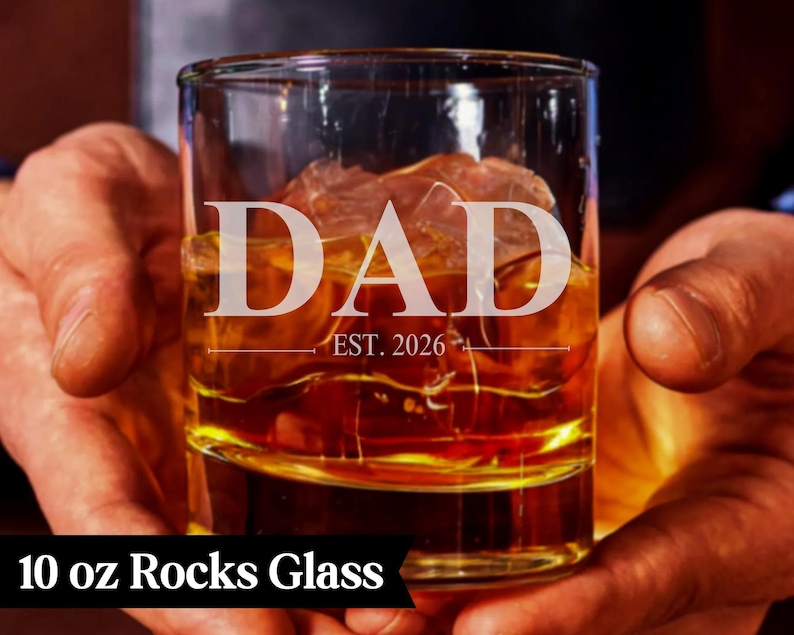 A person's hands holding a 10 oz glass filled with amber-colored liquid, with the word "DAD" and "EST. 1926" engraved on the glass.