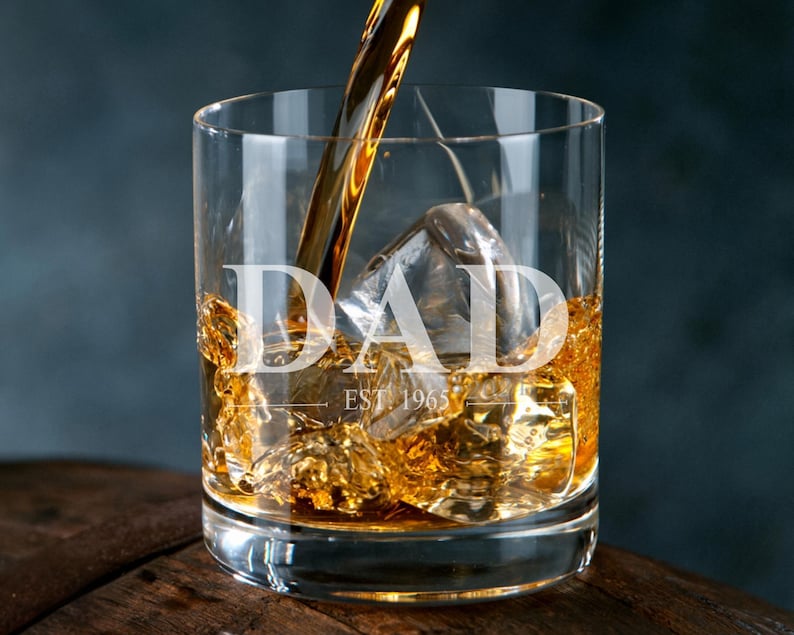 A glass of amber-colored liquid, likely whiskey, with ice cubes, and a golden-colored object being poured into the glass.