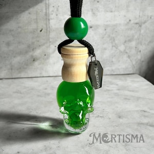 May include: Green skull-shaped glass bottle with a wooden lid and a black cord with a green bead. The bottle has a metal tag that says "Mortisma".