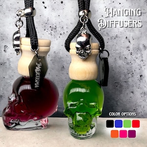 May include: Two hanging skull-shaped glass diffusers with different colored liquids. The diffuser on the left is filled with a dark red liquid and the diffuser on the right is filled with a bright green liquid. The diffusers are hanging from black cords with a silver skull charm. The text "HANGING DIFFUSERS" is at the top of the image. The text "COLOR OPTIONS" is at the bottom of the image with a row of colored squares.