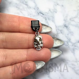 May include: A silver skull charm with a black plastic connector. The charm is hanging from a chain.