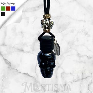 May include: A black skull-shaped car air freshener with a black cord and silver skull accents. The air freshener has a small tag and the word "MORTISMA" is visible at the bottom. Color options are shown in the upper left corner.