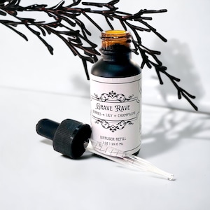 May include: A black glass bottle with a dropper, labeled "Grave Rave Berries + Lily + Champagne Diffuser Refill 1 oz | 29.6 ml".