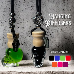 May include: Two hanging car air fresheners shaped like skulls. The air fresheners are black and green and have a wooden top. The air fresheners are hanging from black cords with silver skull beads. The text "HANGING DIFFUSERS" is in the upper right corner of the image. The text "COLOR OPTIONS" is in the lower right corner of the image. Below the text are nine colored squares.