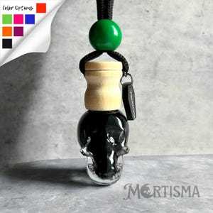 May include: A skull-shaped car air freshener with a black liquid interior. It has a wooden top, a green bead, and a black cord for hanging. The text "MORTISMA" is at the bottom, with color options in the corner.
