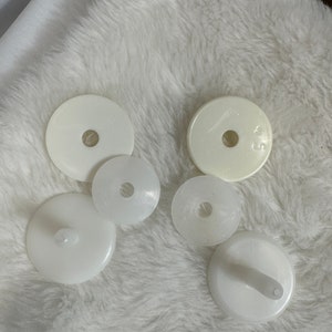Op de afbeelding: Vijf witte plastic schijven met een gat in het midden. Twee van de schijven hebben een verhoogde knop aan de onderkant.