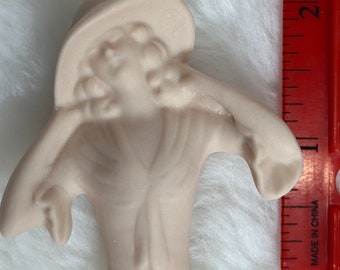 Może przedstawiać: Ceramiczna figurka kobiety w kapeluszu i płaszczu. Figurka jest pomalowana na jasnoróżowy kolor.