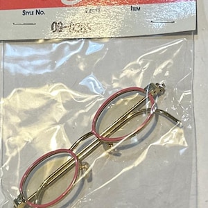 May include: Miniature gold-tone eyeglasses with pink rims, packaged in a clear plastic bag. The glasses have oval lenses and delicate temples. The packaging includes the text "La Sioux" and "Style No. 09-0335".