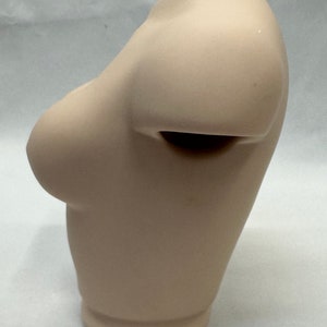 May include: A beige ceramic bust sculpture with a smooth, rounded surface. The bust is missing the head and neck, and the shoulders are sculpted in a relaxed pose.